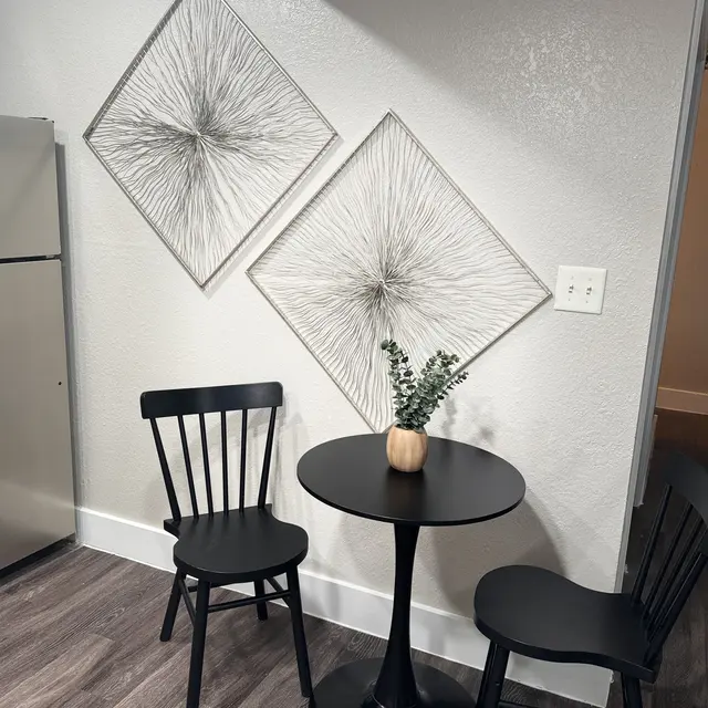 A modern kitchen nook featuring a small black round table with two black chairs. A decorative vase with greenery sits on the table. Above the table, two diamond-shaped wall decorations with a radial design are mounted. The walls and flooring are light-colored, creating a sleek and inviting atmosphere.