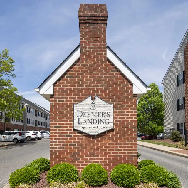 A brick sign for Deemer's Landing apartment complex, surrounded by green shrubs and trees, with buildings in the background.