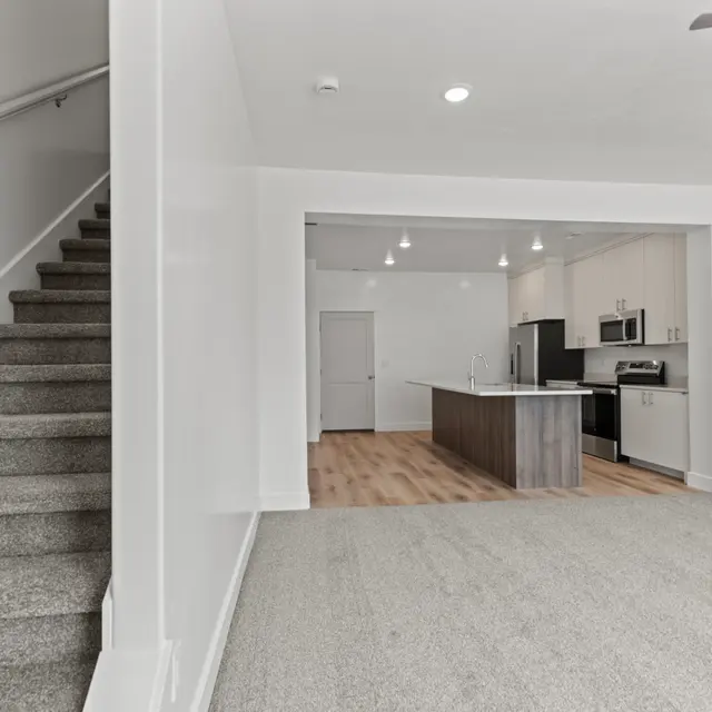A spacious modern living area with a kitchen visible in the background. A staircase leads to an upper level, and the floor is carpeted while the kitchen features a countertop island.