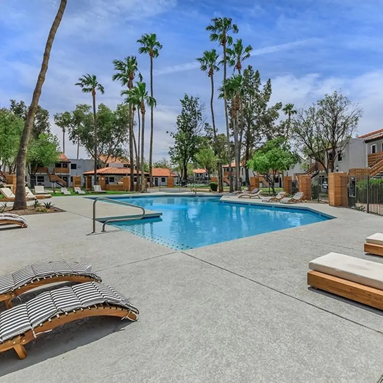 A serene residential pool area featuring a clear blue swimming pool surrounded by lounge chairs, palm trees, and landscaped greenery. The pool is bordered by apartment buildings with a warm color scheme.