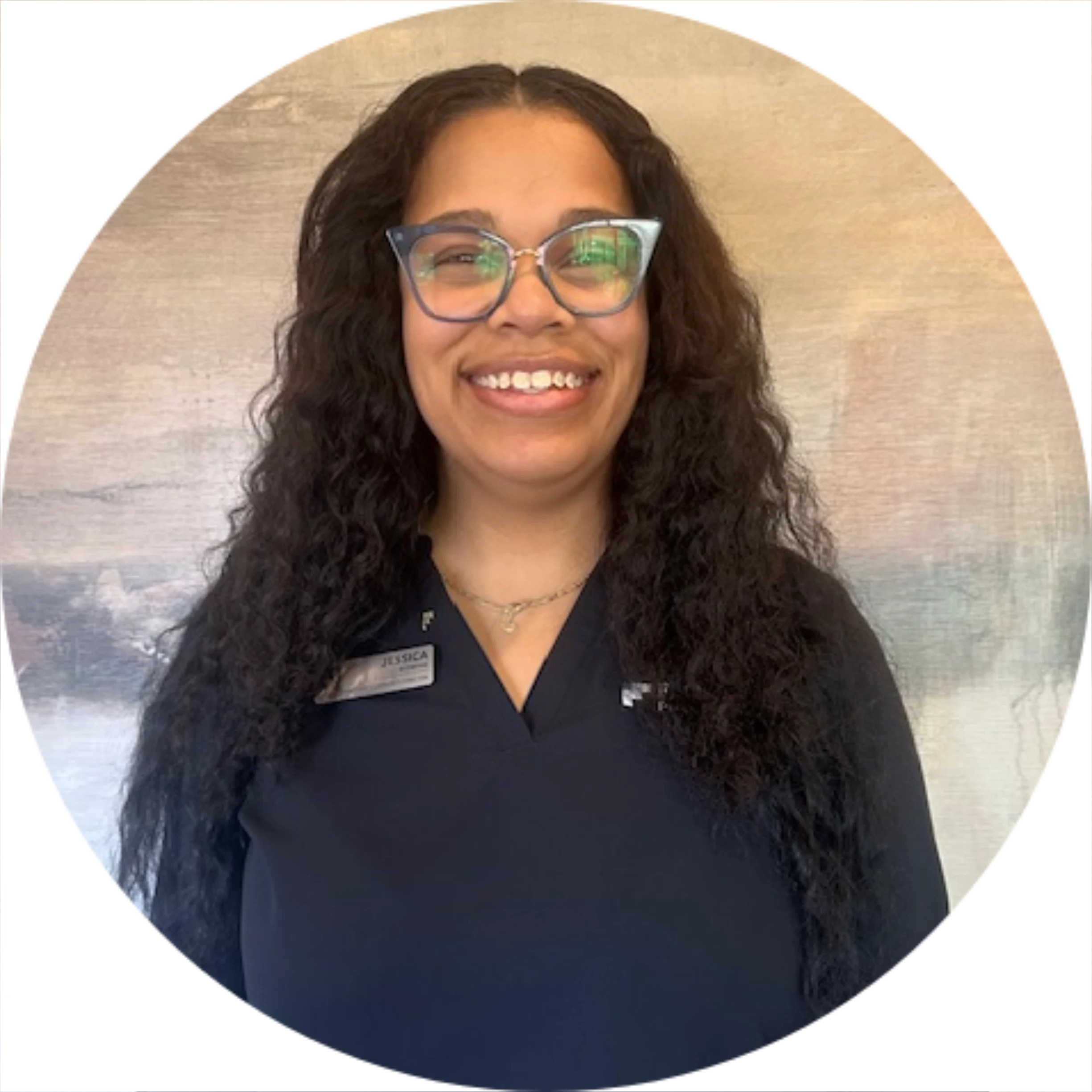 A smiling woman with long curly hair wearing glasses and a dark uniform, standing against a light textured background.