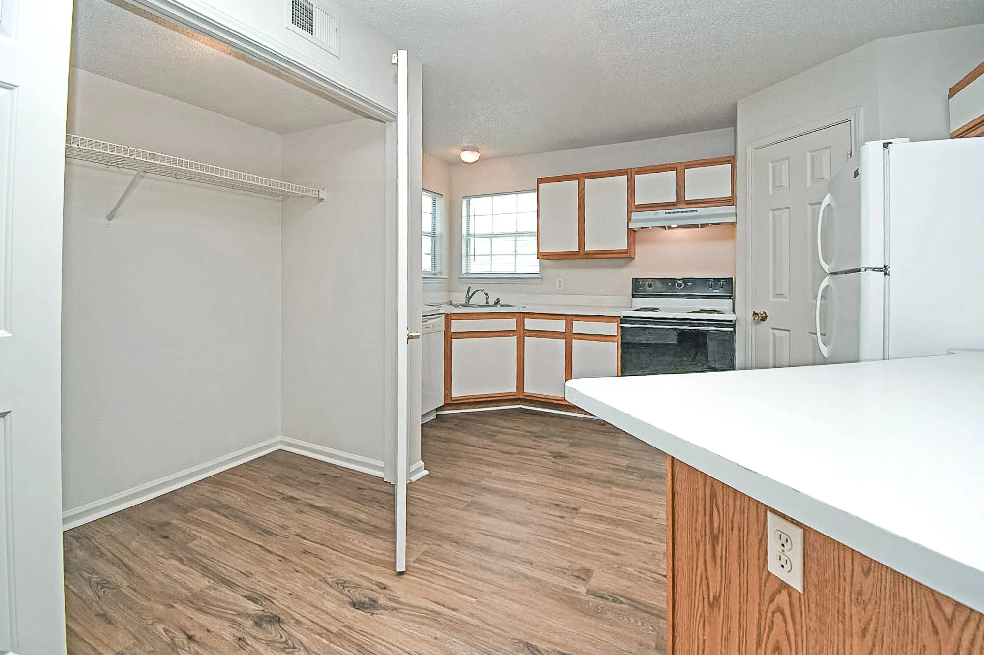 A view of a kitchen area with white cabinets, a stove, a sink, and a spacious closet to the side. The floor is wooden, and there is ample natural light coming in from a window.