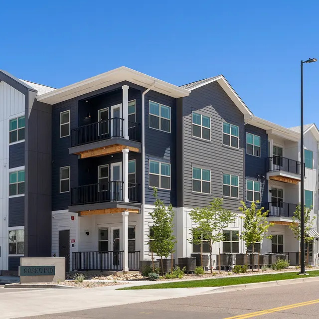 A modern apartment complex featuring multiple stories with a contemporary design, including balconies and lush green landscaping in front.