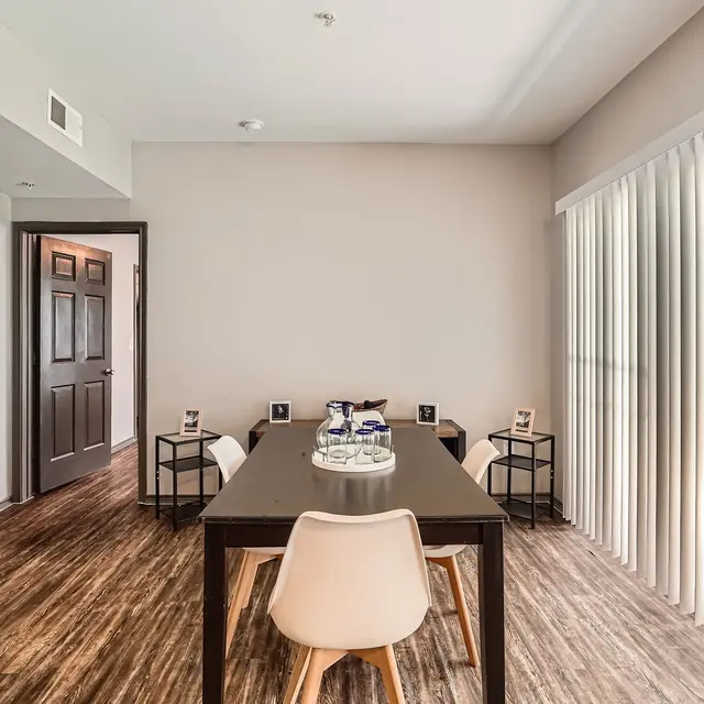 A modern dining area featuring a wooden table with white chairs, surrounded by large windows with vertical blinds, and a neutral color palette on the walls.