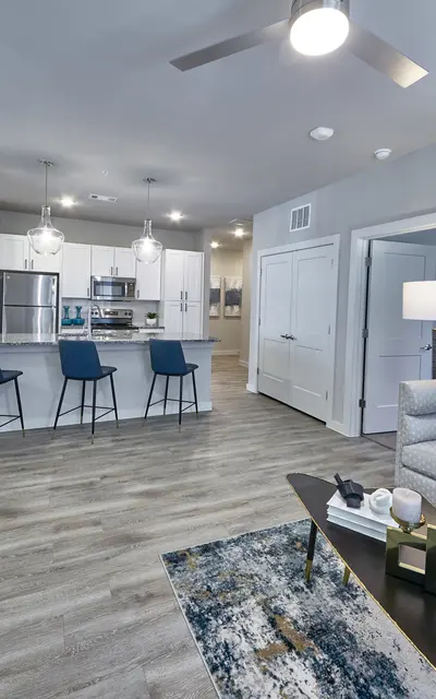 A modern living room with a gray sofa, decorative pillows, and a stylish coffee table. A kitchen with a large island and bar stools can be seen in the background.