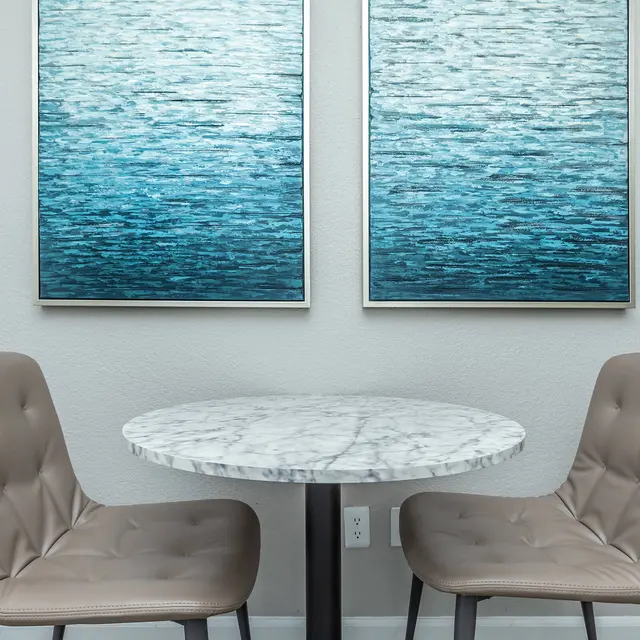 A modern dining setup featuring a marble tabletop and two beige upholstered chairs, accompanied by two abstract blue art pieces on the wall.