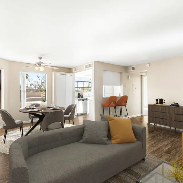 A modern living space featuring a neutral color palette, a gray sofa, an orange accent chair, and a circular dining table with four chairs. Large windows allow natural light, and there are decorative elements like a small plant and stylish furniture.