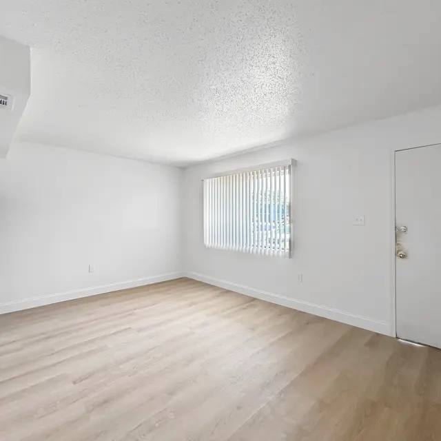 Bright Empty Room Interior An empty room with light wood flooring, a refrigerator on the left, and a window with vertical blinds letting in natural light. There is a white door on the right leading outside.