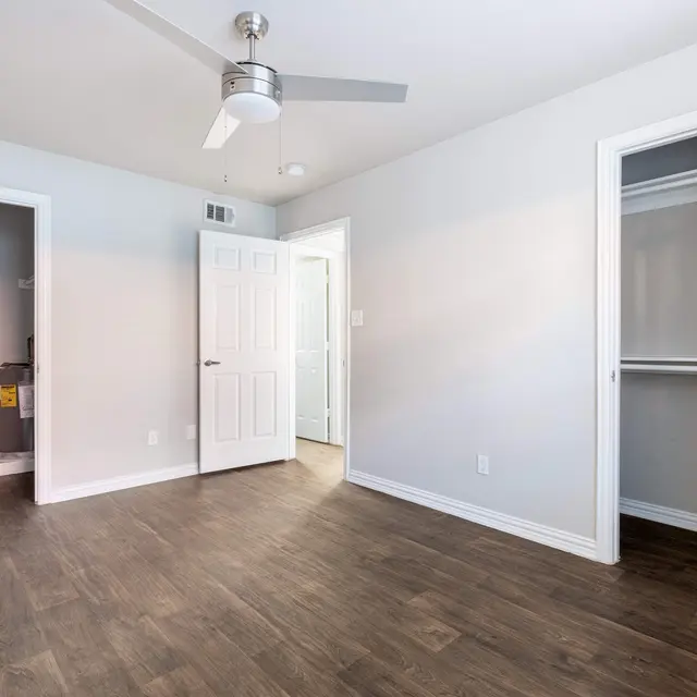 An empty bedroom with light gray walls and wood flooring. Two open doors on either side lead to adjacent rooms, and a closet door is visible on the right. A ceiling fan hangs in the center of the room.