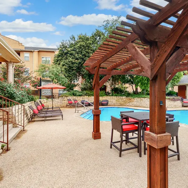 A swimming pool area with a pergola, lounge chairs, and landscaping. The pool is surrounded by a textured surface and there are buildings in the background with trees.