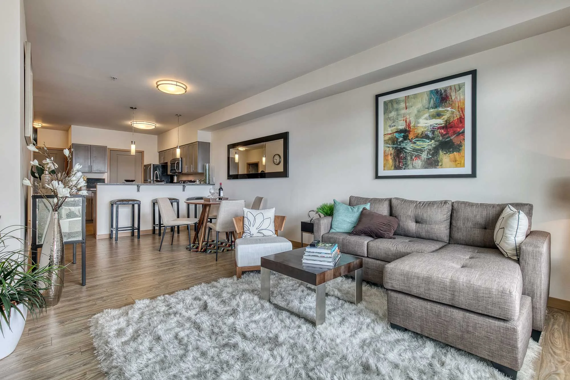 A modern living room with a cozy sectional sofa, a coffee table, and an area rug. The space features light wood flooring and a large artwork on the wall. In the background, there's a dining area and a kitchen with modern fixtures.