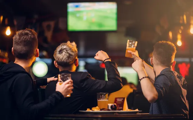 Three friends watching a sports game on a large screen in a bar, holding drinks and cheering excitedly.
