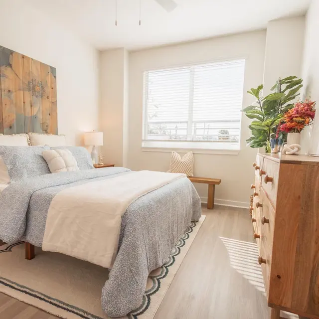 A cozy bedroom featuring a queen-sized bed with light blue bedding, a decorative wall art with floral design, a wooden dresser, a potted plant, and a window letting in natural light.