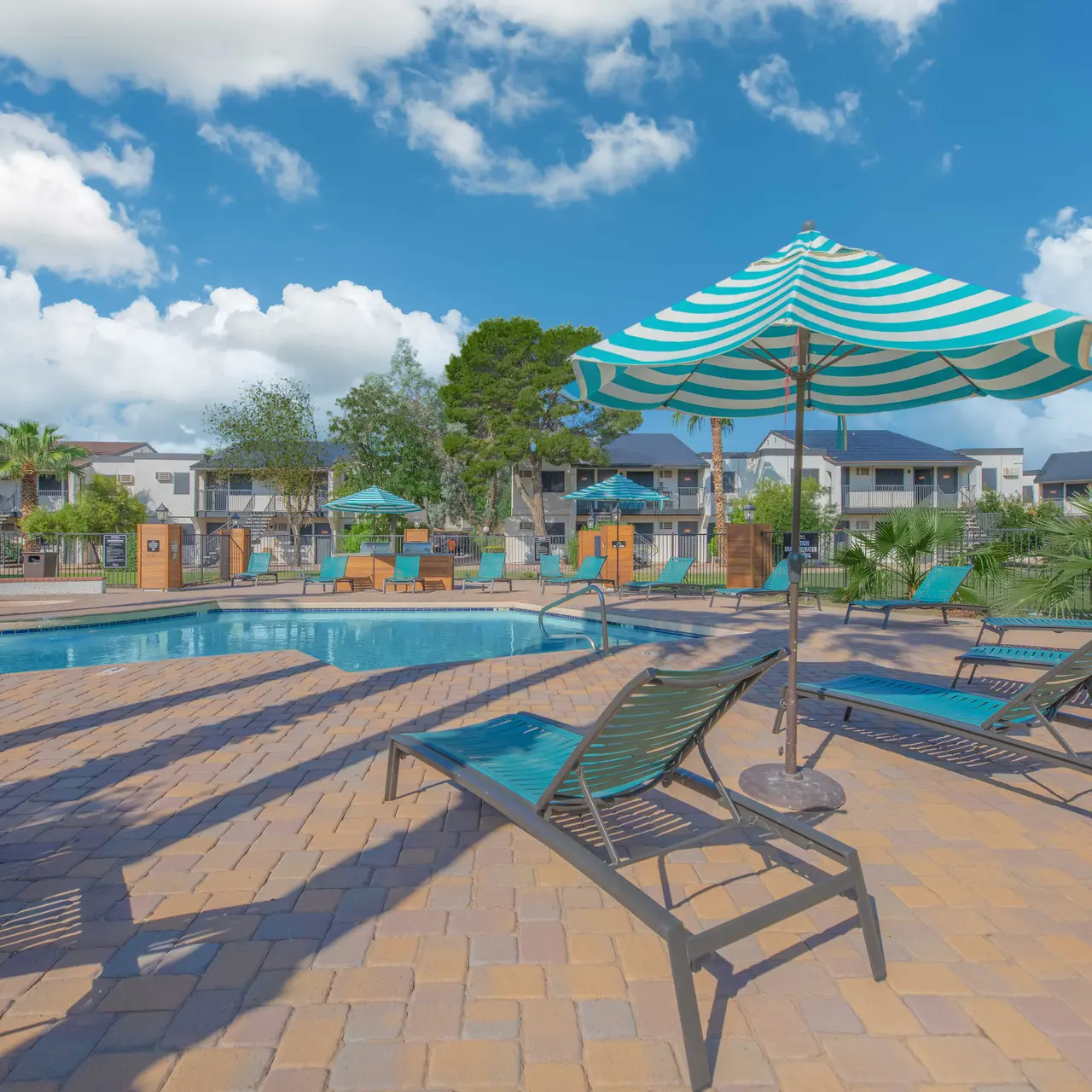 A sunny swimming pool area featuring lounge chairs and a striped umbrella. The pool is surrounded by greenery and modern buildings.