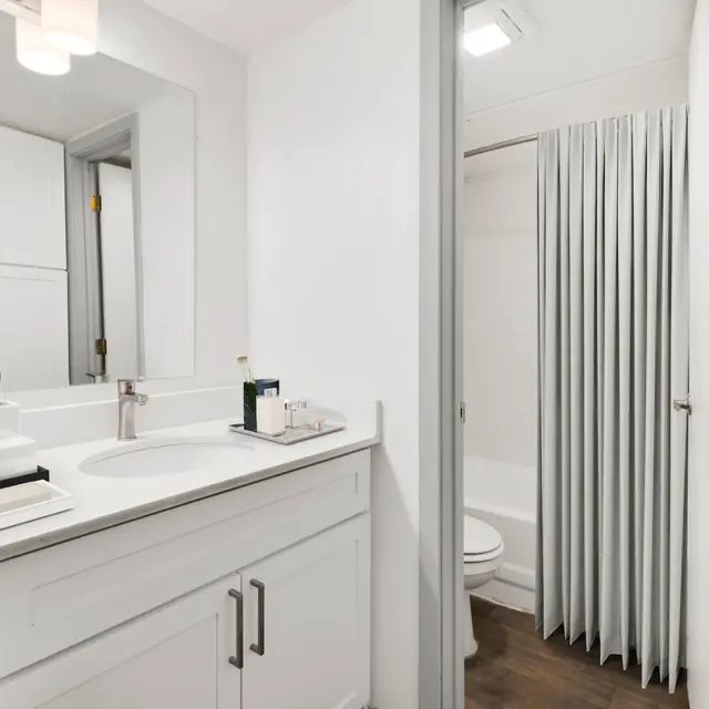 A modern bathroom with a white vanity, round mirror, and a shower with a curtain. The space features light walls, a wooden floor, and decorative items on the vanity.