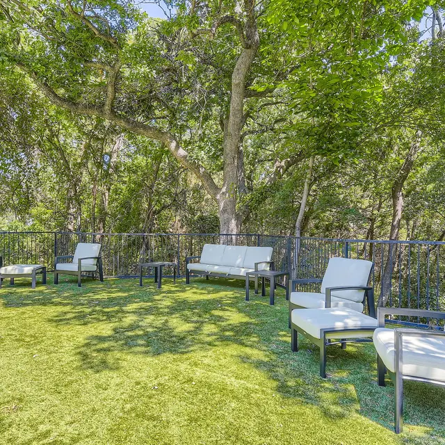 An outdoor seating area surrounded by trees, featuring white chairs on a green grass-like surface.