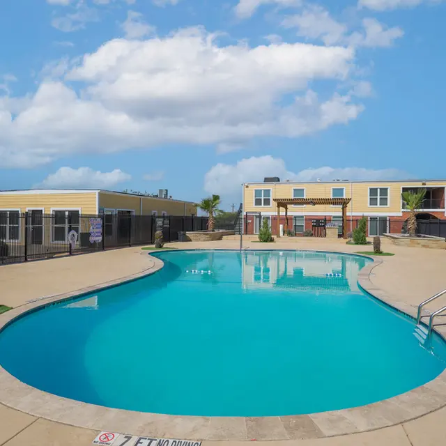 A large swimming pool surrounded by a paved area, with palm trees and lounge chairs. In the background, there are two-story apartment buildings and a blue sky with fluffy clouds.