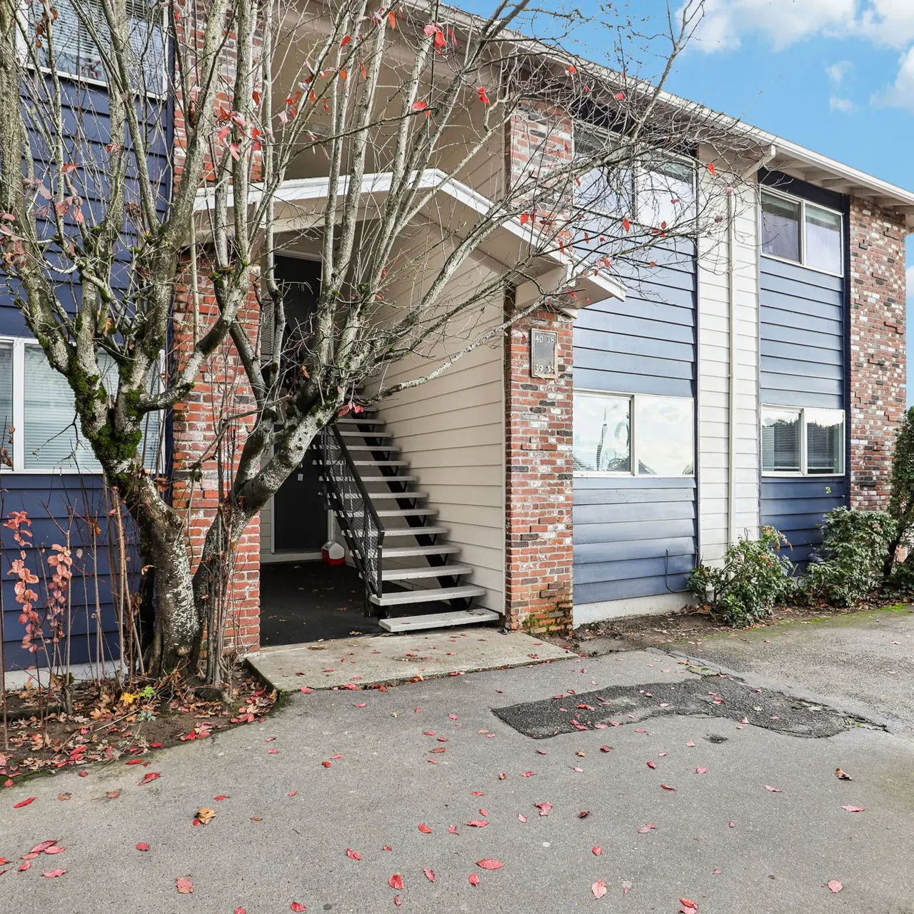 Exterior view of a two-story apartment building with blue and brick siding, showcasing a tree in front and a walkway leading to a set of stairs.