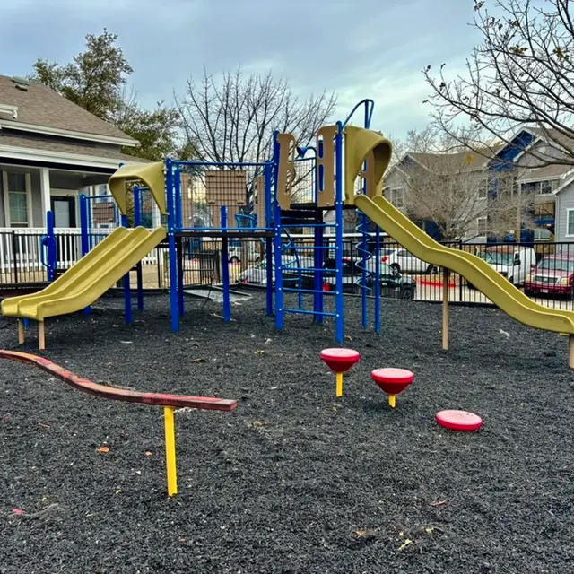 A playground featuring two yellow slides, climbing structures, and a balance beam with red circular platforms, set in a black mulch area.