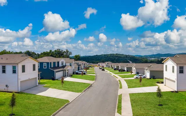 A scenic view of a residential neighborhood with rows of houses on either side of a paved road, green lawns, and a backdrop of hills under a blue sky with fluffy clouds.