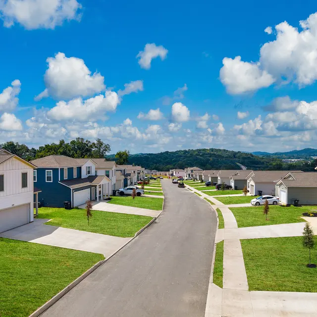 A scenic view of a residential neighborhood with rows of houses on either side of a paved road, green lawns, and a backdrop of hills under a blue sky with fluffy clouds.