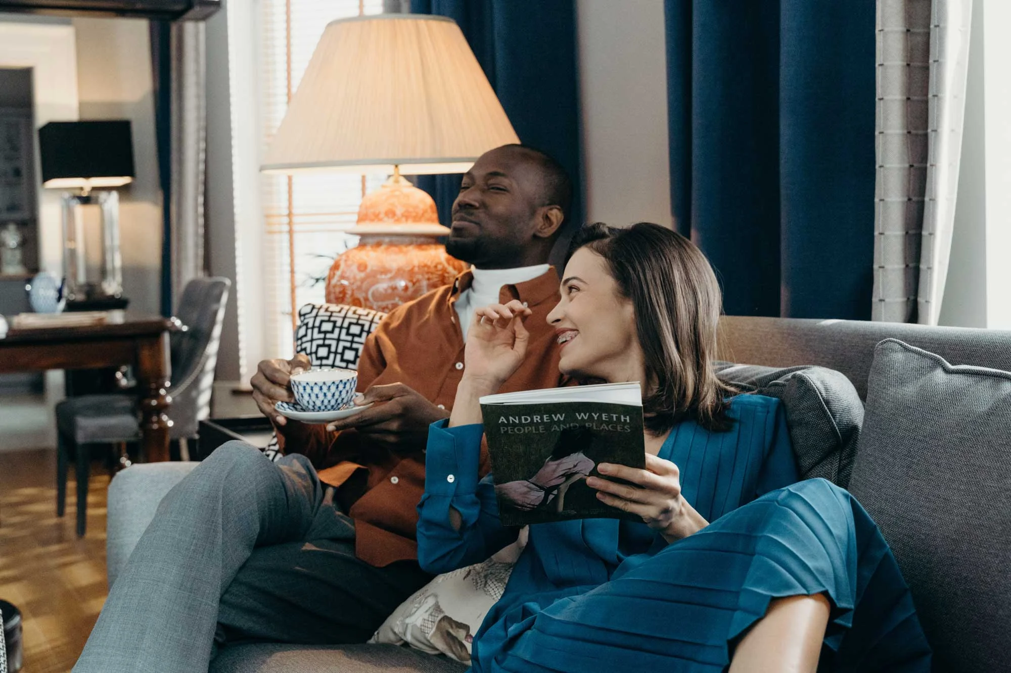 A man and a woman sitting together on a couch, sharing a moment in a cozy living room. The woman holds a book, while the man enjoys a cup of tea. They both appear engaged and happy.