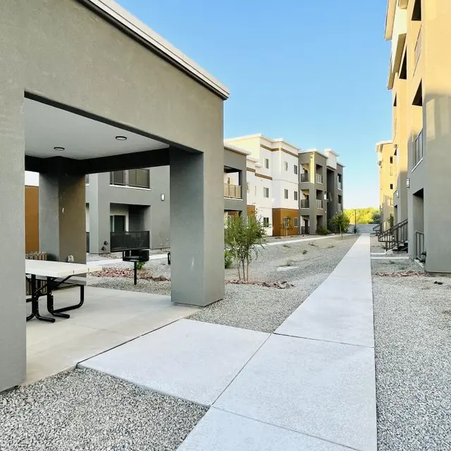 A modern apartment complex featuring grey buildings with a landscaped pathway. There are picnic tables under a covered area and some green plants along the pathway.