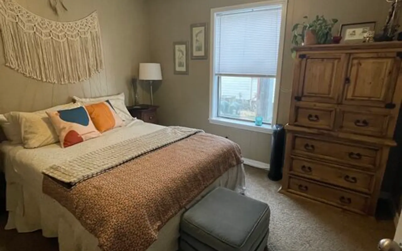 Cozy Bedroom Interior A cozy bedroom featuring a double bed with decorative pillows, a bedside lamp, a window with blinds, a wooden dresser, and a macrame wall hanging.