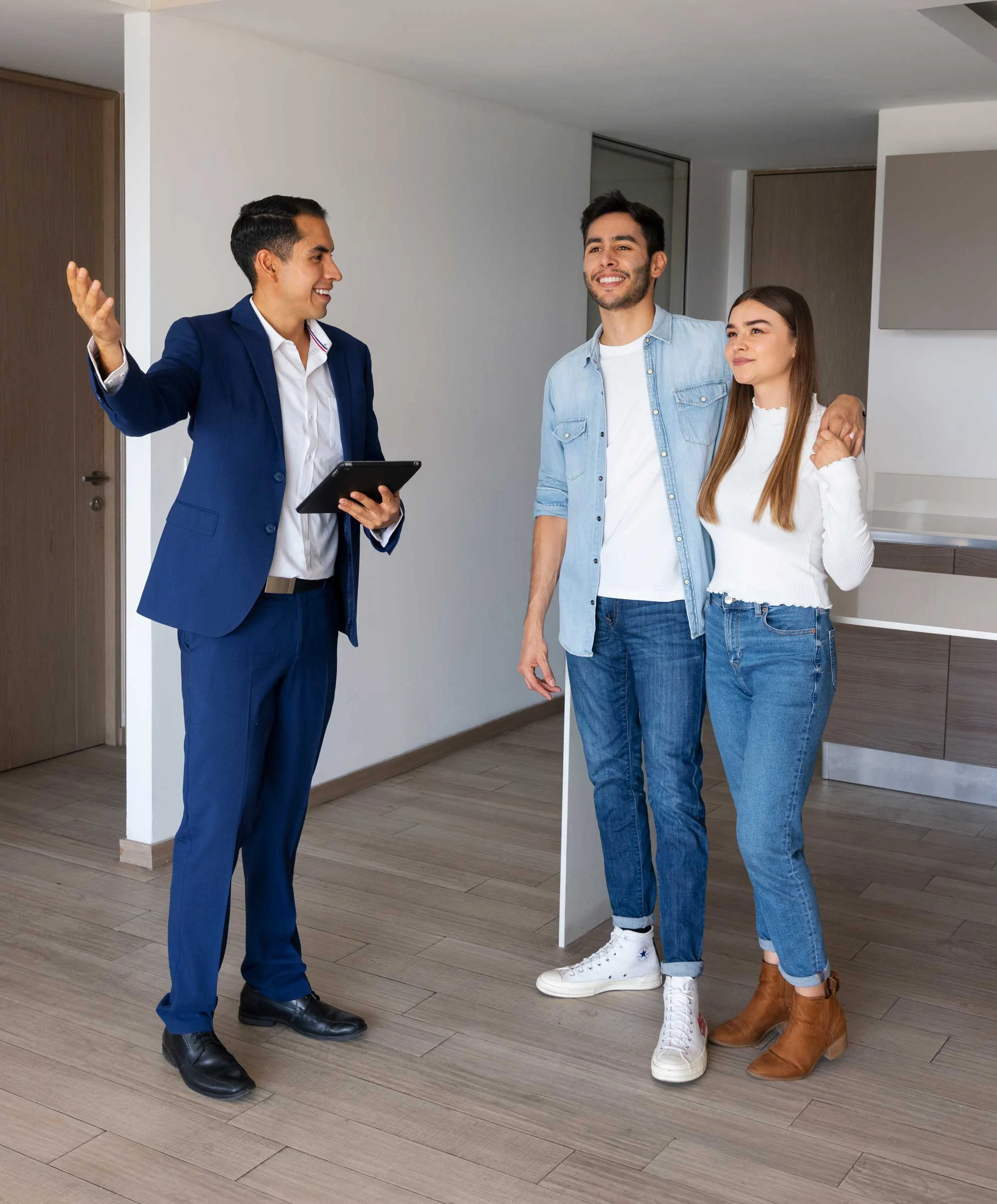 A real estate agent showing a modern apartment to a young couple. The agent is smiling and gesturing, while the couple appears to be engaged and interested in the space.