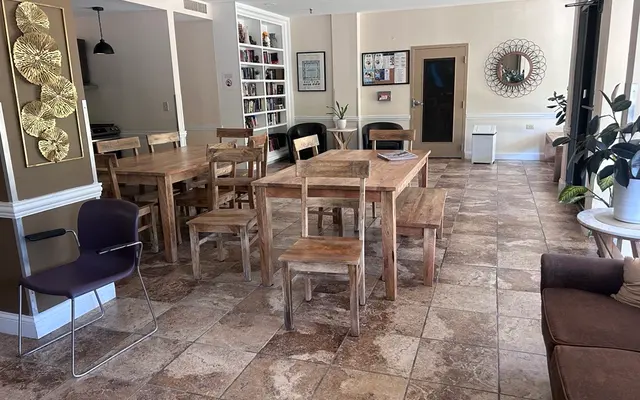 Community Gathering Space A spacious community room featuring wooden dining tables and chairs, a bookshelf, plants, and ample natural light from large windows.