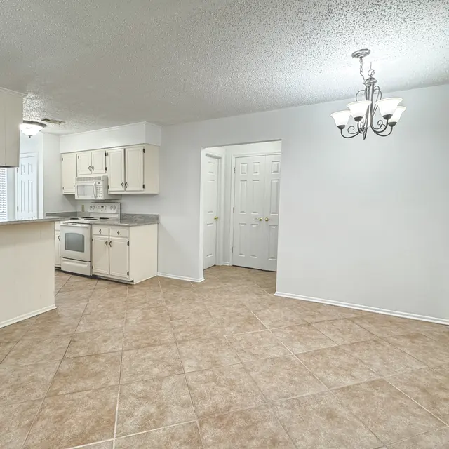 A spacious kitchen and dining area with light-colored walls and tile flooring. The kitchen features light cabinets, a stove, and a view to the dining area below a chandelier.
