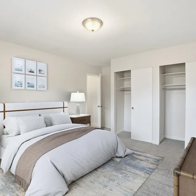 A modern and spacious bedroom featuring a bed with neutral bedding, two bedside tables with lamps, and a decorative rug. There are two empty closet doors and artwork on the wall.