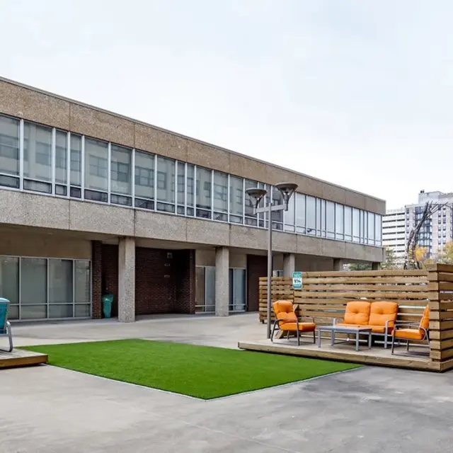 An outdoor area with modern furniture, featuring orange and teal armchairs surrounded by wooden partitions and green grass-like turf, adjacent to a large building.