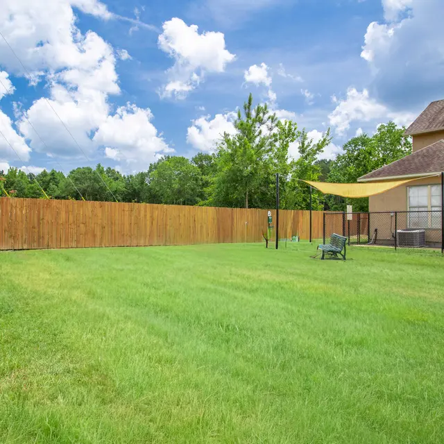 A spacious backyard with green grass and a wooden fence. There are trees in the background and a building on the right side.
