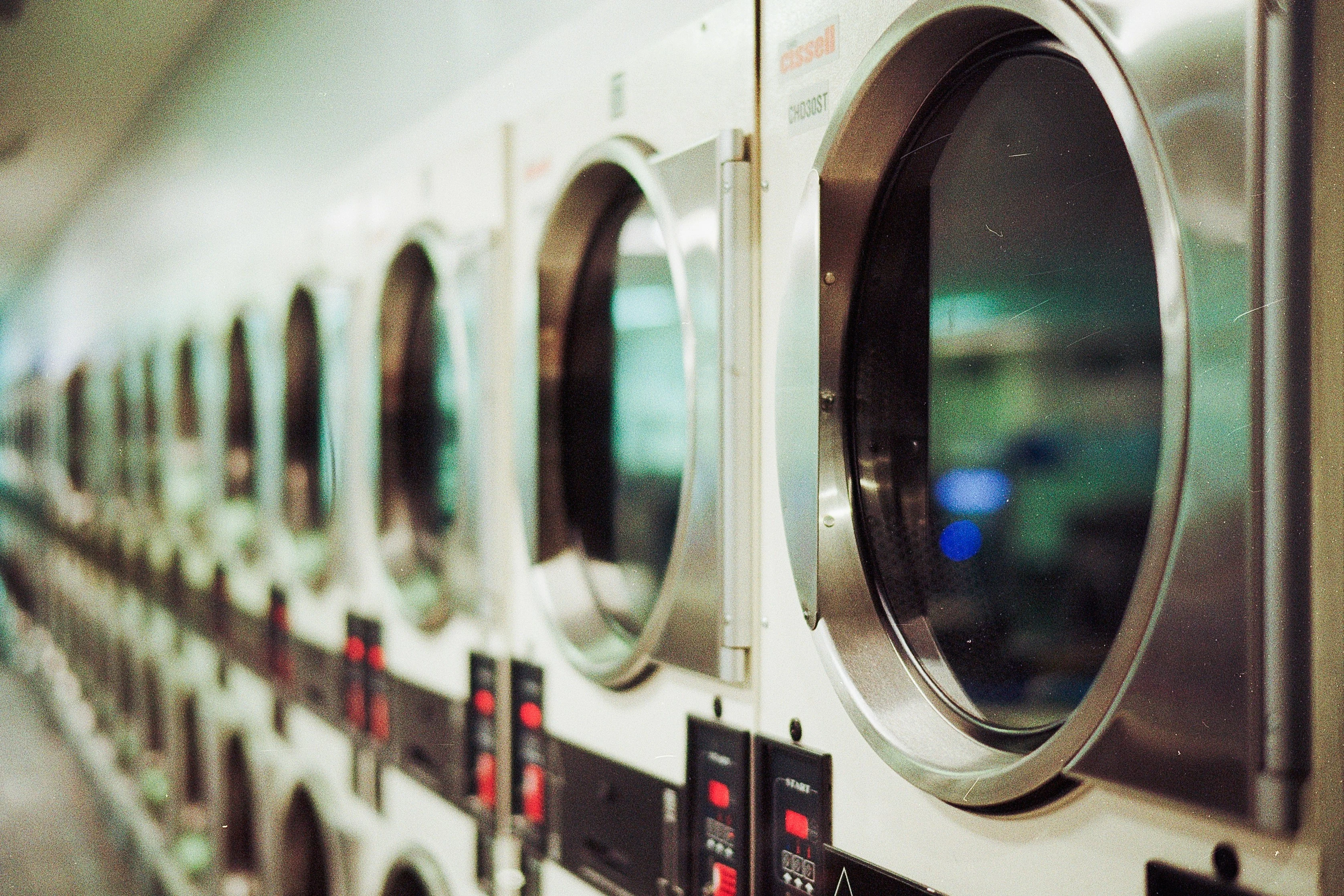 A row of washing machines with round glass doors, showcasing a sleek and modern design. The machines have metallic exteriors and are lined up against a wall, with control panels below each door.