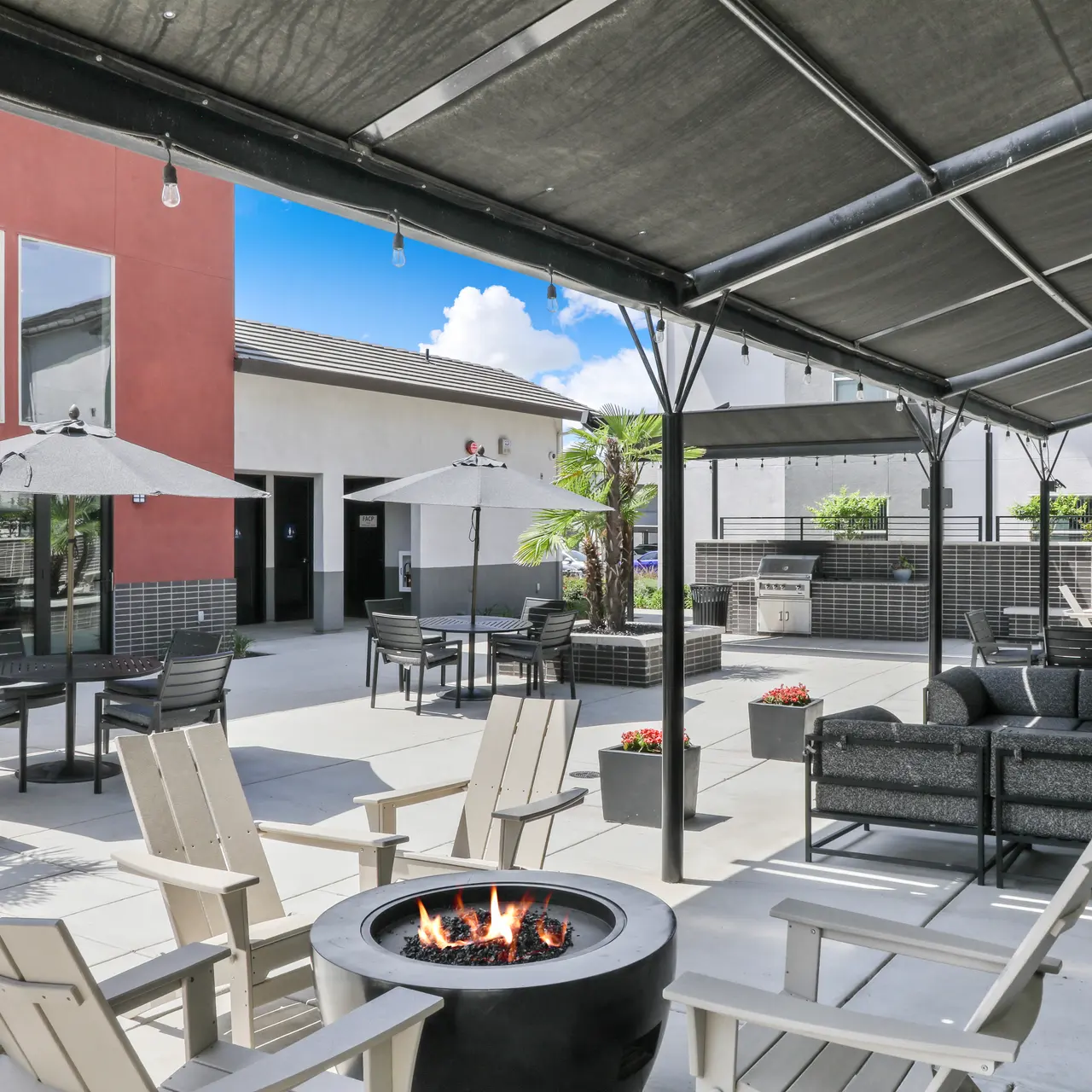 A modern outdoor lounge area featuring a fire pit, comfortable seating, umbrellas, and lush greenery.