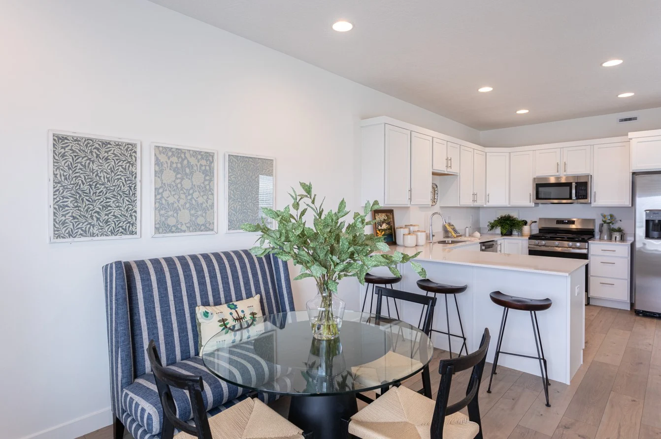 A modern kitchen and dining area with a round black glass table and striped upholstered sofa. Potted plant on the table. White cabinetry and stainless steel appliances are visible in the kitchen background.