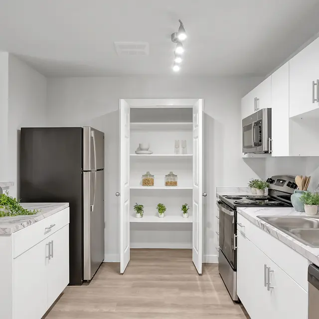 A modern kitchen featuring white cabinets, stainless steel appliances, and a pantry with open shelves. The room has a light and airy feel with natural wood flooring.