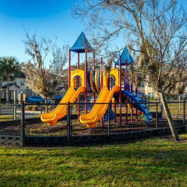 Colorful playground equipment featuring yellow and blue slides surrounded by a fenced area with grass and nearby houses.