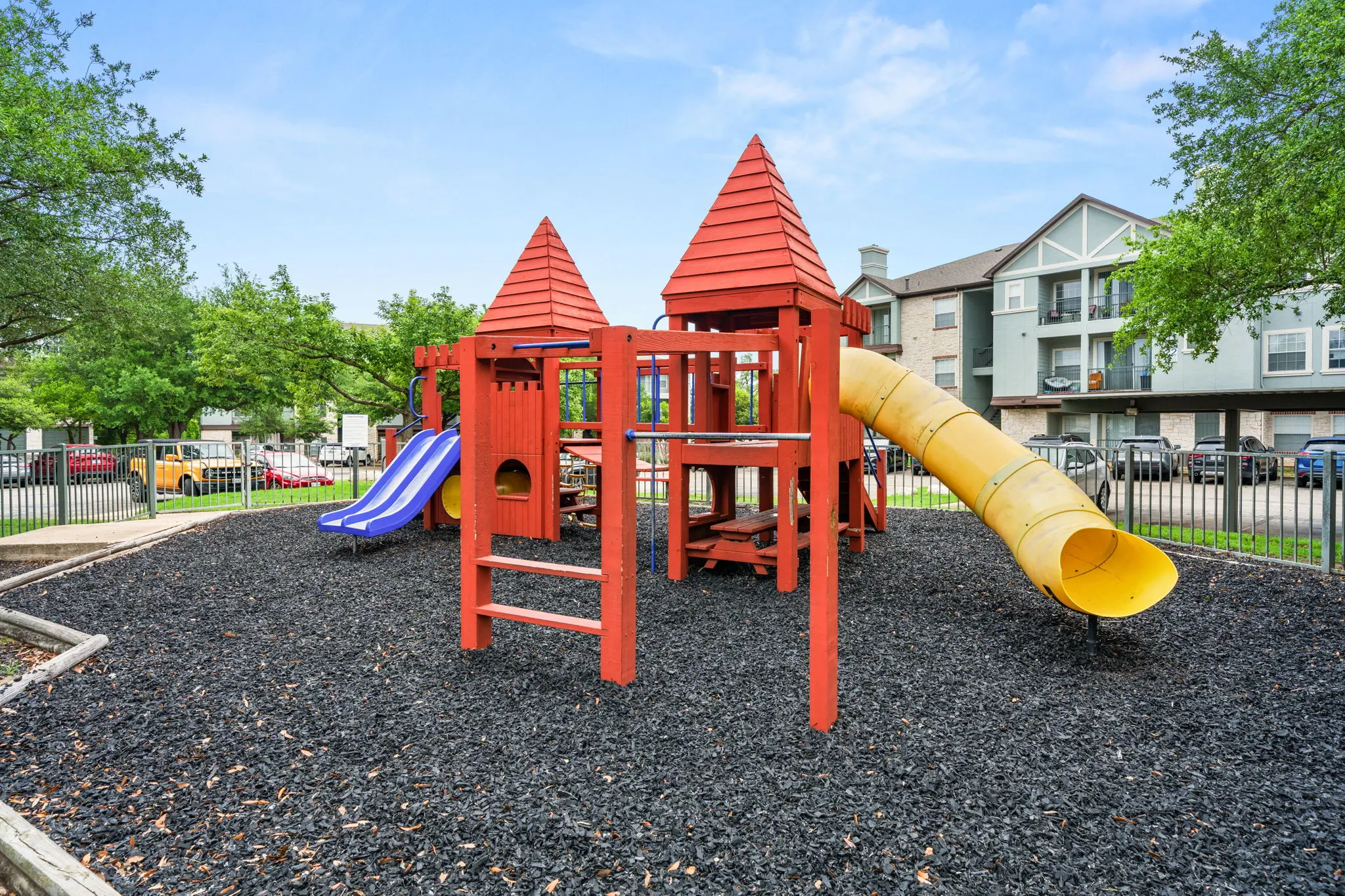 Brightly colored playground structure with red towers, a yellow slide, and a blue slide on black rubber mulch in a park setting.