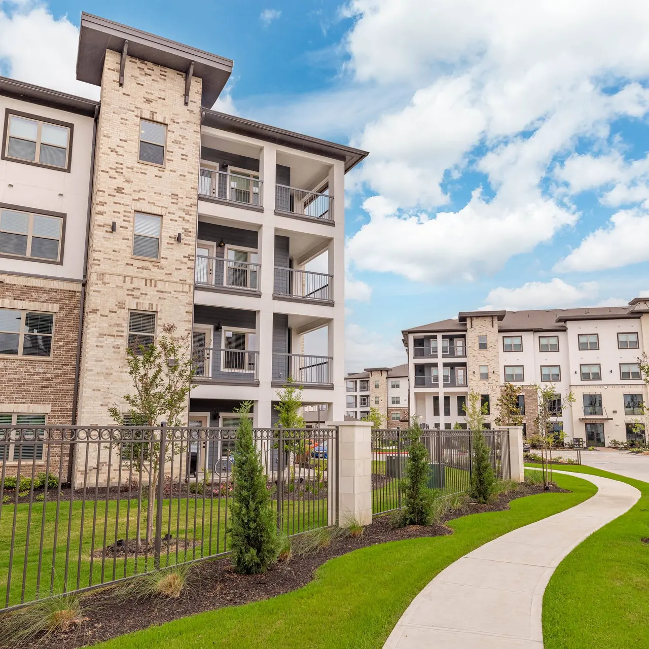 A modern multi-story apartment complex with stone and brick facade, surrounded by green lawns and neatly trimmed shrubs. The sky is blue with a few clouds.