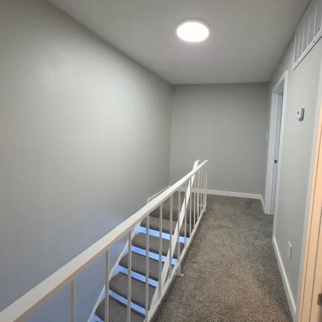 A well-lit interior hallway with a white railing leading down a staircase, gray walls, carpeted floor, and doors on the right side.