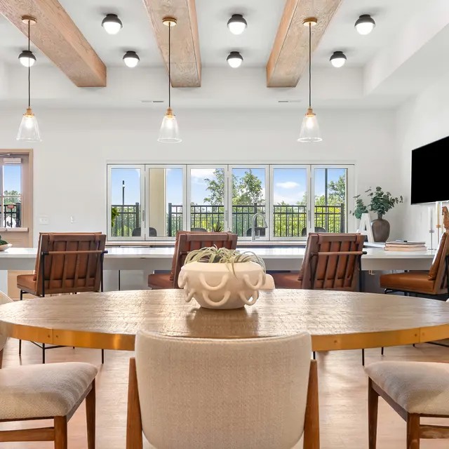 A modern dining area featuring a round wooden table with beige upholstered chairs. There are four wooden chairs with brown cushions positioned around the table. The room has large windows providing natural light, with greenery visible outside. The ceiling has exposed wooden beams and recessed lighting fixtures. A wall-mounted TV and a small plant add decor to the space.