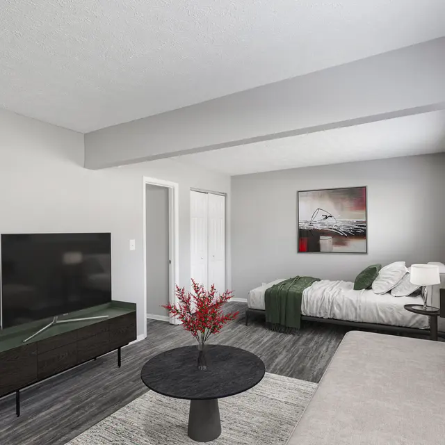 The Hideaway A modern living space with a gray painted room, featuring a black flat-screen TV on a stylish TV stand, a round black coffee table with a decorative plant, a beige sofa, and a made bed with green and white bedding. A piece of artwork hangs on the wall, and there are white closets in the background.