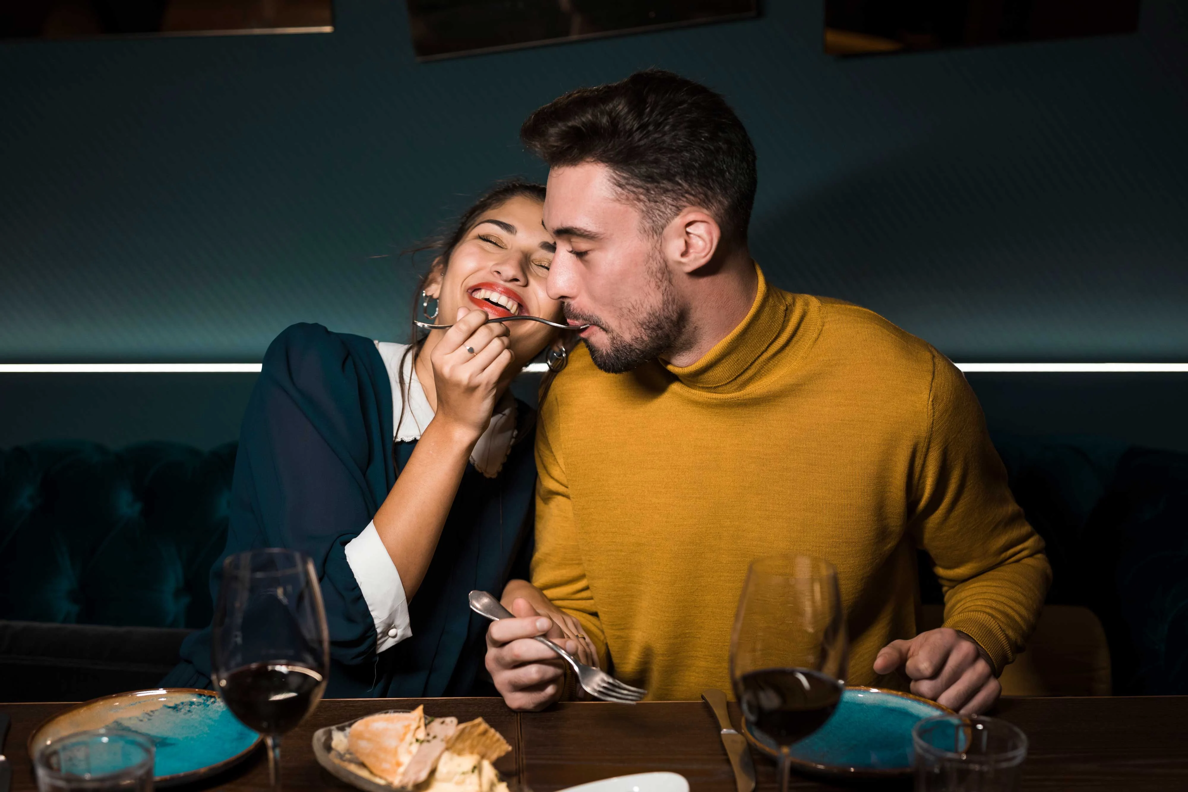 A couple enjoying a playful moment at dinner in a cozy restaurant setting. The woman is laughing as she playfully touches the man's face, while he looks amused. They are surrounded by plates of food and wine glasses.