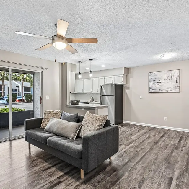 Modern Living Room Design A modern living room with a dark gray sofa, light wooden floors, and a ceiling fan. The room features large sliding glass doors that lead to a patio, and an open kitchen area visible in the background.