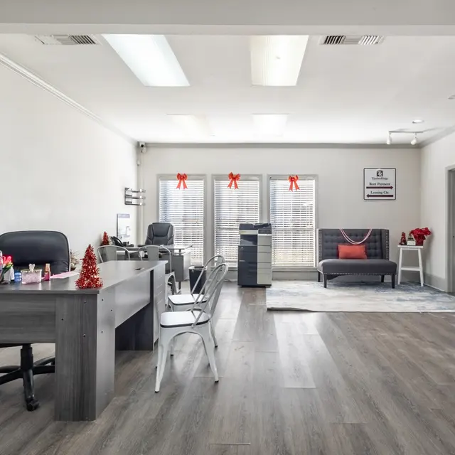 A modern office interior featuring a desk with a computer, a chair, and decorative items. There is a cozy seating area with a gray couch and festive decorations visible through large windows.