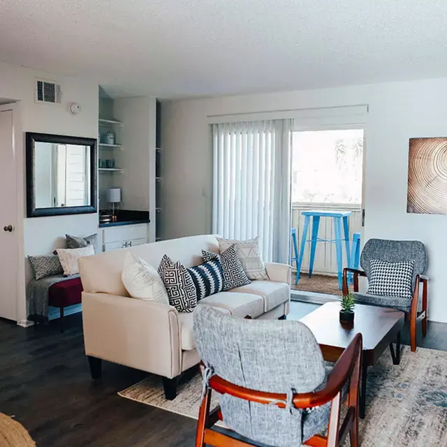 A modern living room featuring a light-colored sofa, patterned throw pillows, and a wooden coffee table. There are two armchairs and a sliding glass door leading to a balcony. A piece of modern art hangs on the wall.