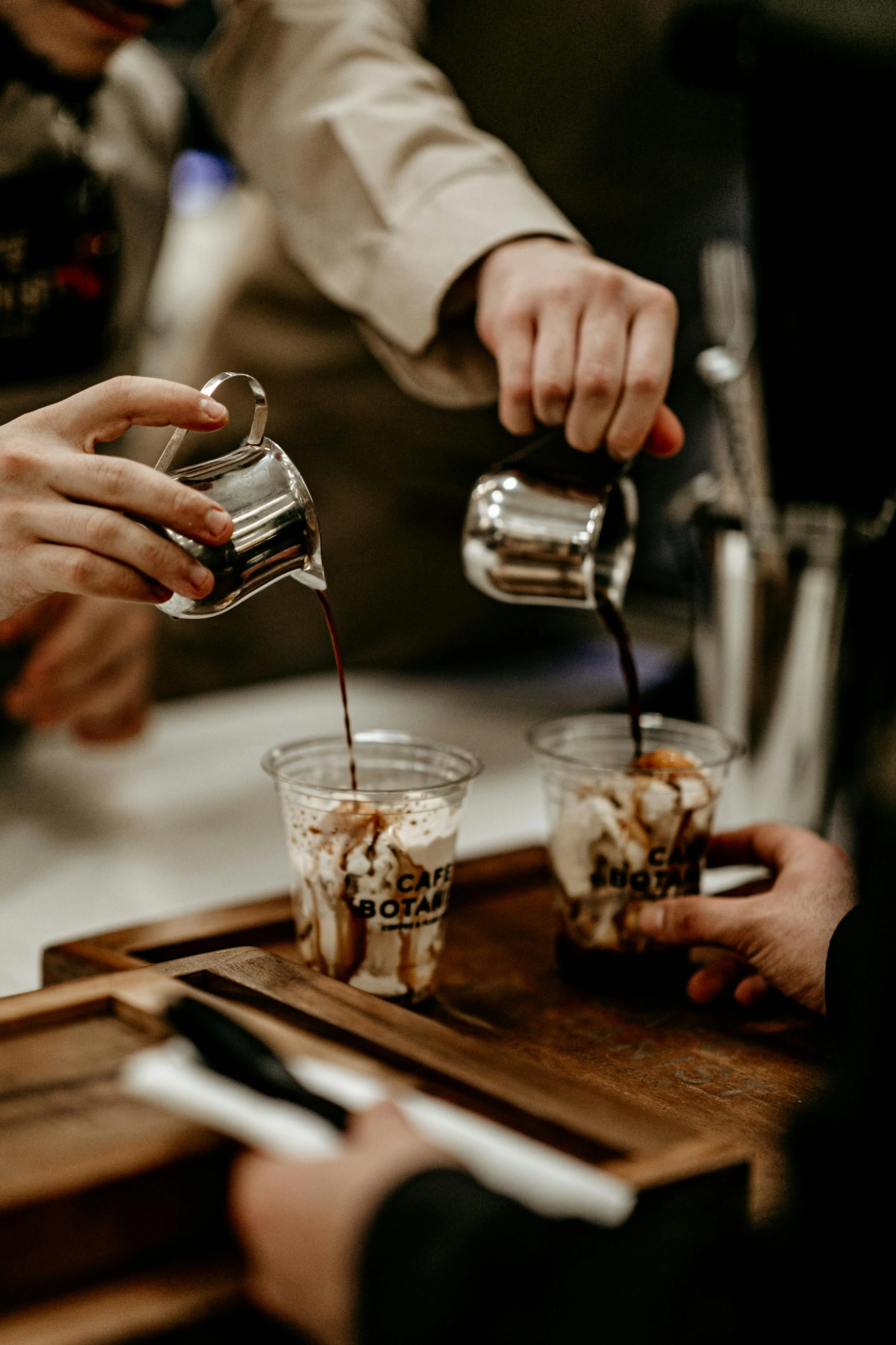 Two hands pouring dark liquid from silver pitchers into clear cups with whipped cream and syrup, placed on a wooden tray.