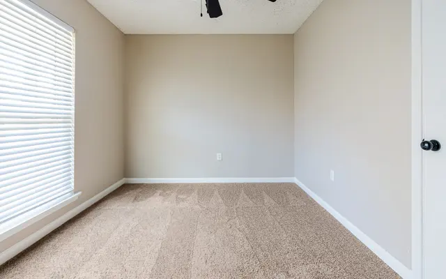 An empty room featuring light beige walls, tan carpet, and a ceiling fan with three blades. A window with blinds allows light to enter the space.